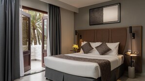 A modern hotel room with a large bed, a side table with a lamp, and a view of palm trees through the open door. - Marbella Resort Sharjah (Sharjah)