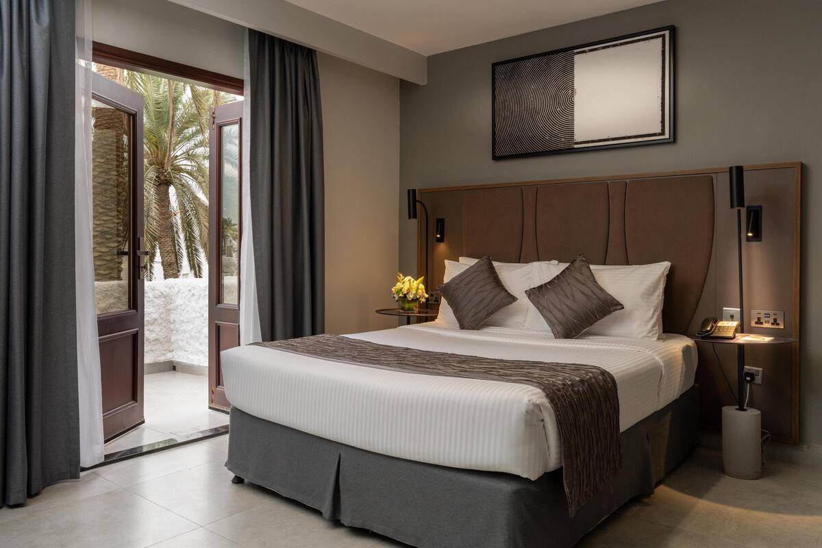 A modern hotel room with a large bed, a side table with a lamp, and a view of palm trees through the open door.