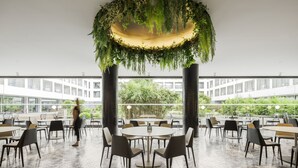 Un espacio interior amplio con mesas y sillas redondas, un jardín en el techo y vistas a un edificio y zonas verdes exteriores.