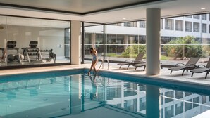 Una piscina interior moderna con paredes de vidrio transparente, un área de descanso junto a la piscina y vista a un edificio en el fondo.