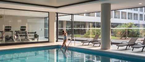 Relax by the pool with a view of the adjacent building through large windows, surrounded by lounge chairs and a seating area.
