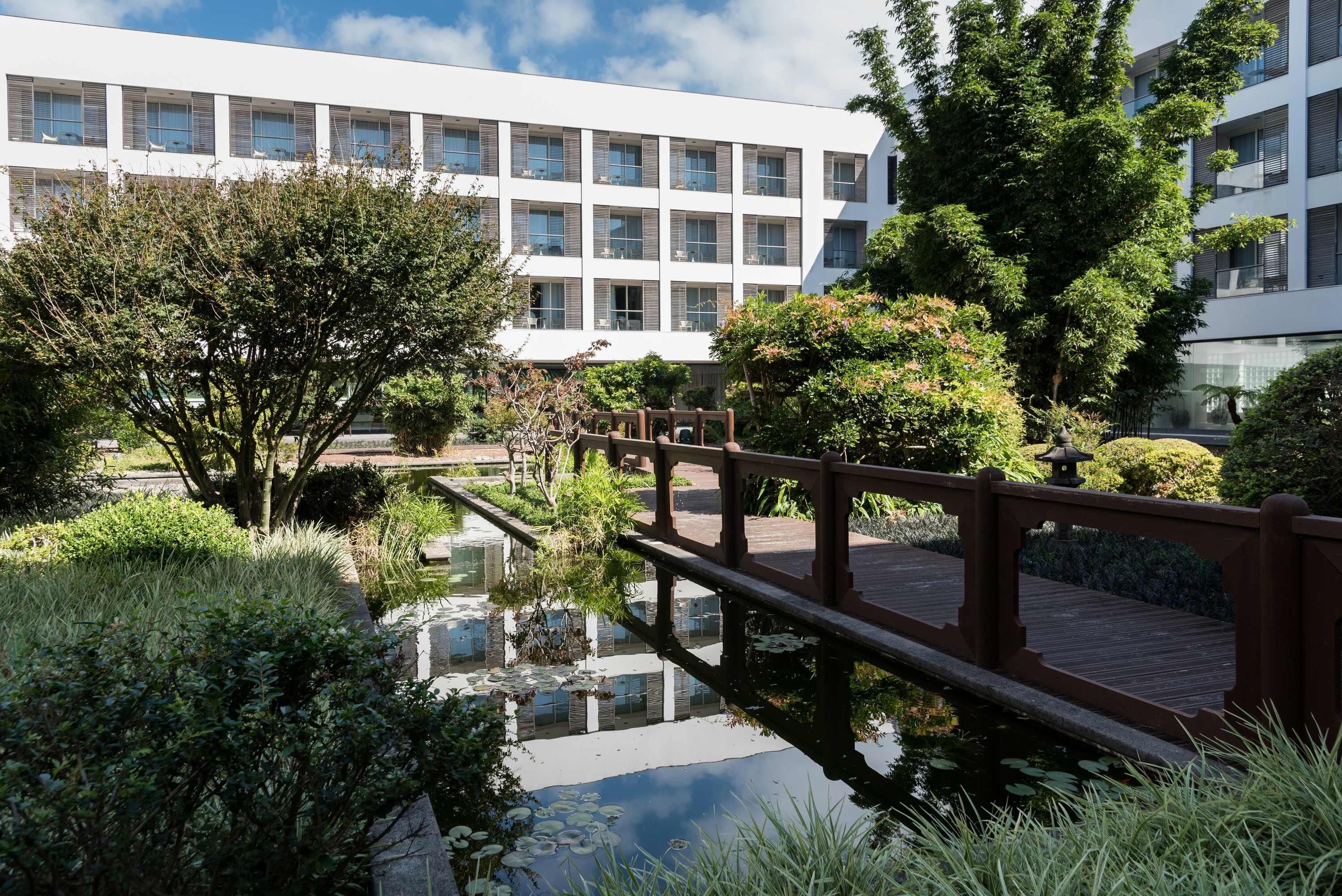 Un bâtiment moderne agrémenté d’un élément décoratif aquatique réfléchissant, d’un pont en bois et d’une végétation luxuriante.