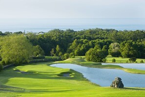 A golf course with a pond, surrounded by dense trees and a view of the ocean.