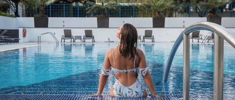 A woman sitting by a pool with a tiled floor, surrounded by chairs and a blue fence.