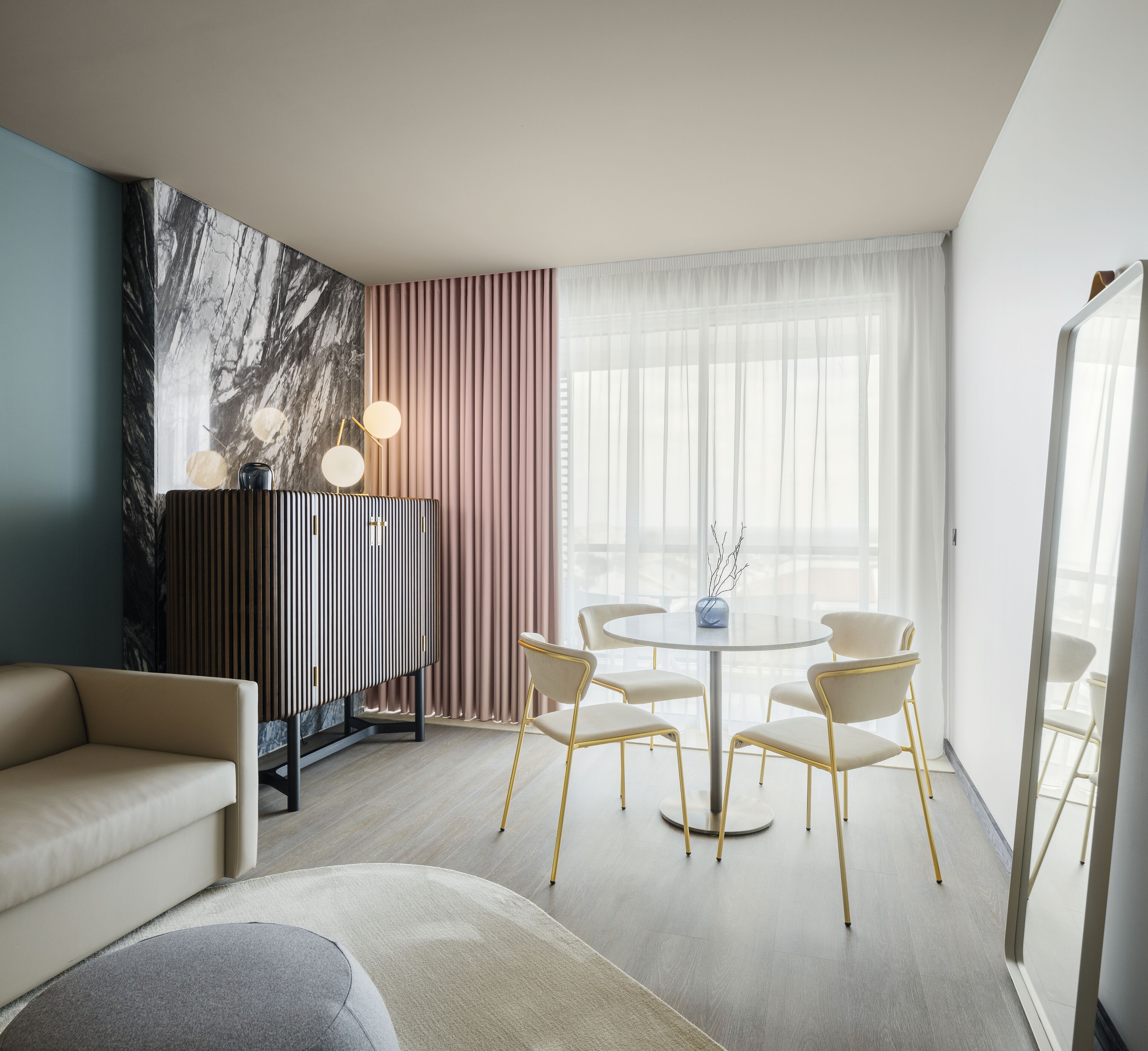 Una habitación de hotel moderna con una pared decorada con mármol, una mesa redonda con sillas, un sofá y un espejo grande.