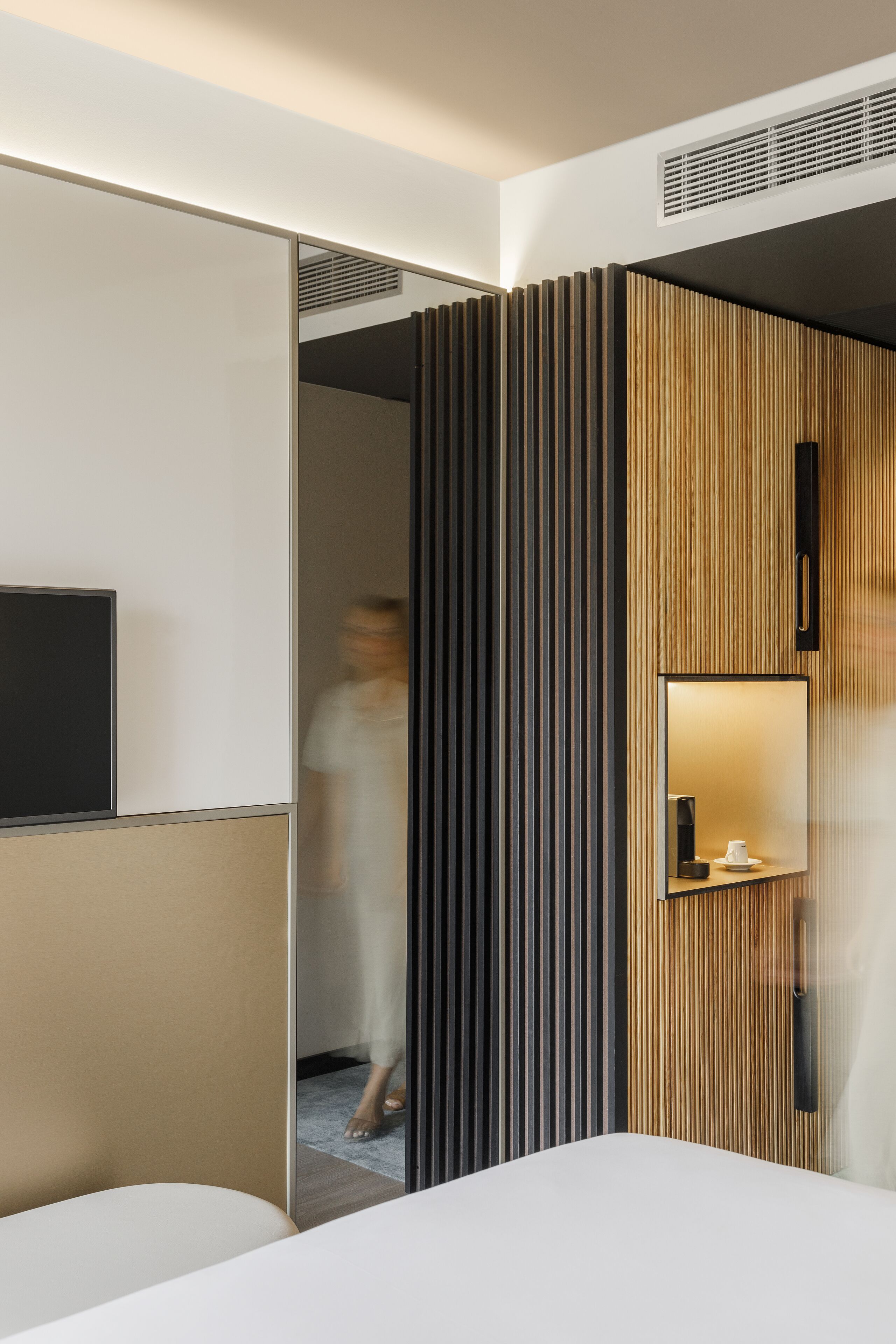 A modern hotel room with a large bed, a bedside table, a mirror, and a TV.