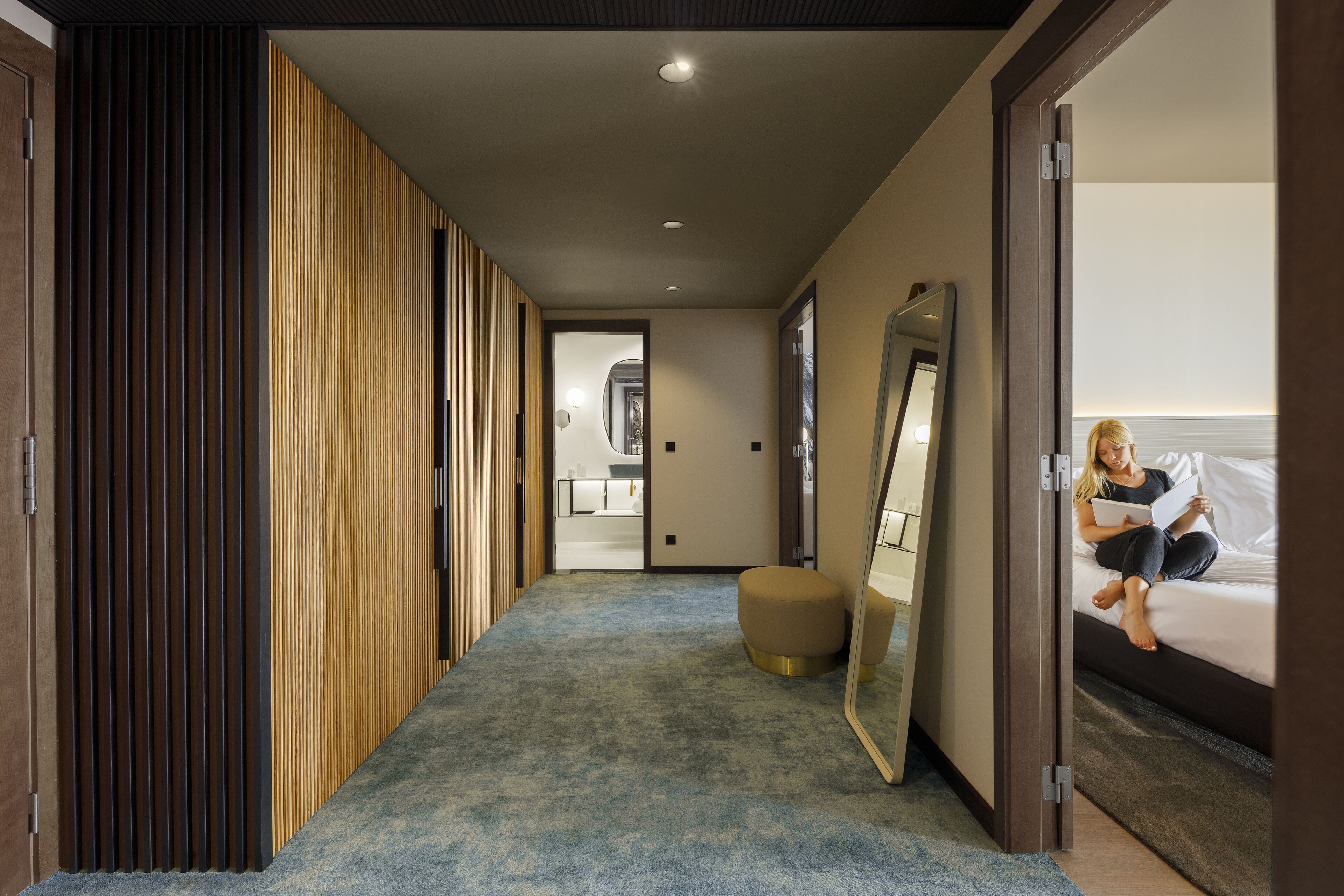 Un pasillo moderno de hotel con un revestimiento de panel de madera, alfombra azul, un otoman redonda y una cama con una persona leyendo un libro.