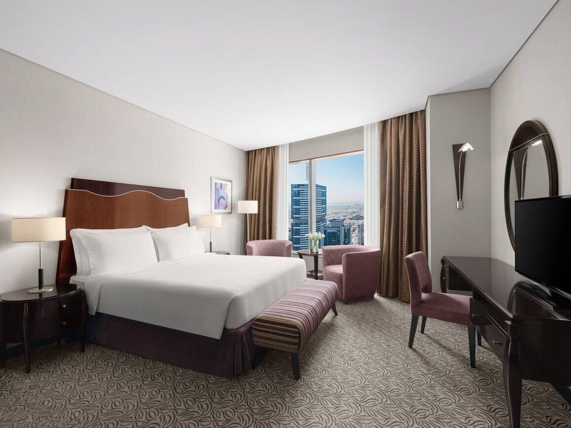 A hotel room with a large bed, a desk, a chair, a TV, and a view of the cityscape.