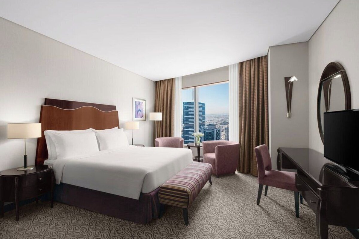 A hotel room with a large bed, a desk, a chair, a TV, and a view of the cityscape.