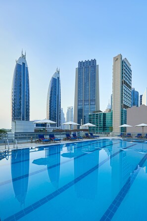 Una piscina con sillas de descanso y sombrillas, rodeada de modernos edificios altos.