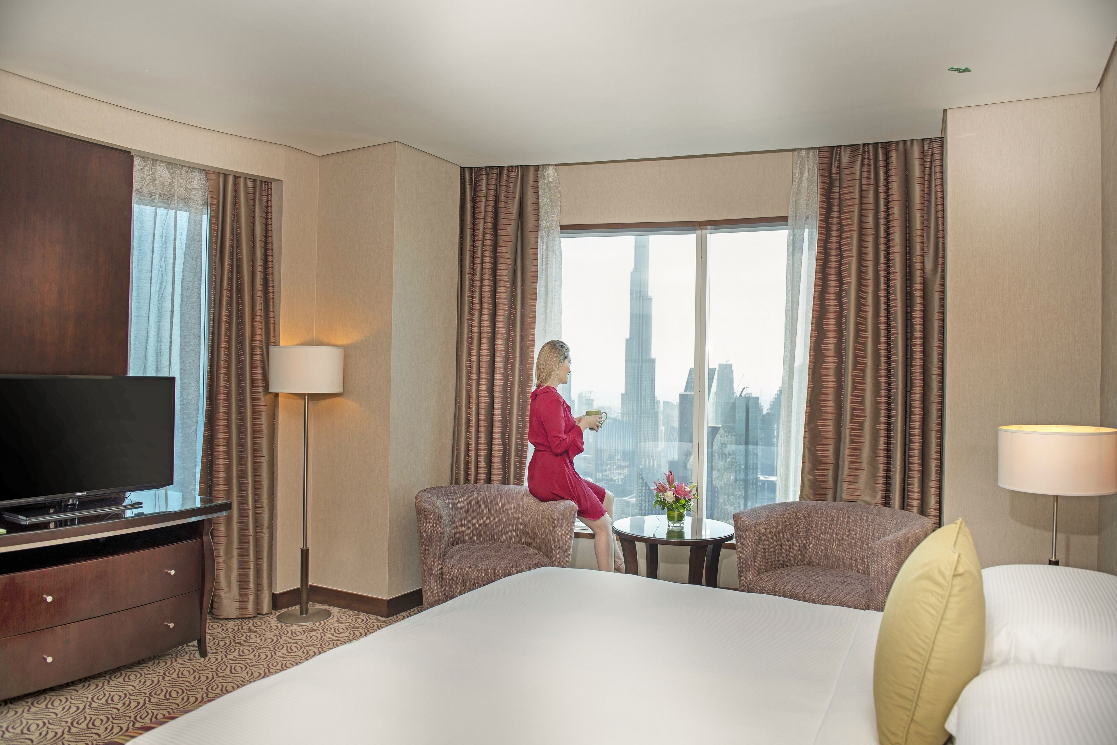 A hotel room with a large bed, two armchairs, a TV, and a window with a city view.