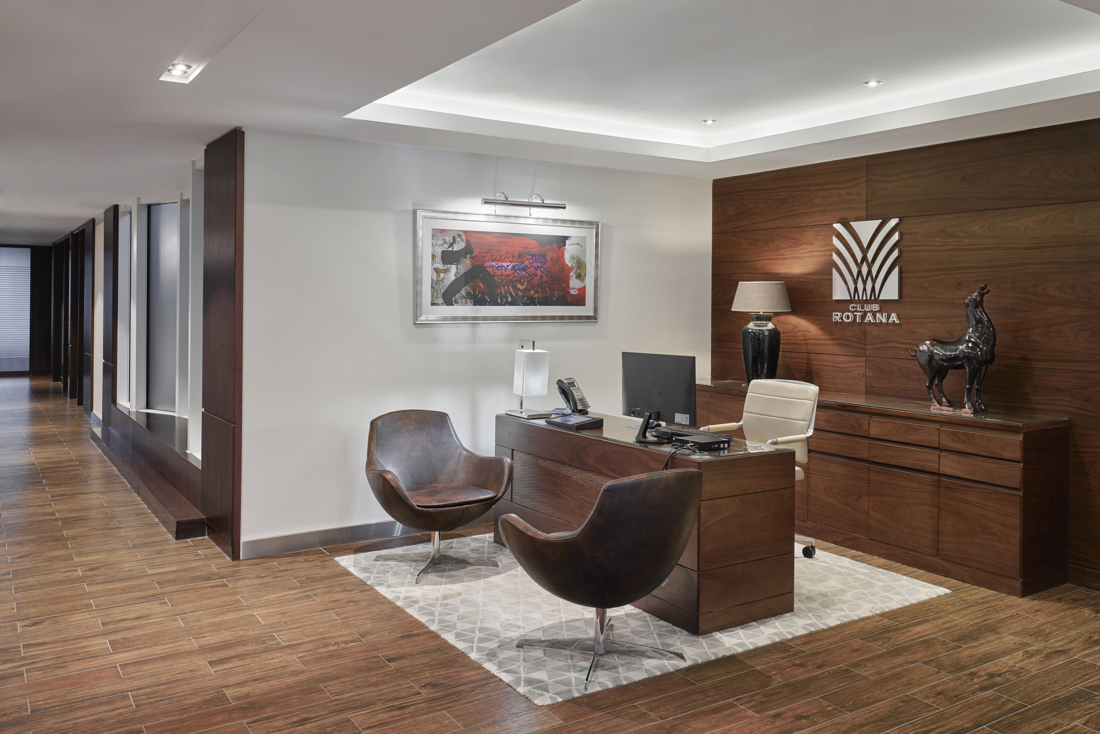 A modern reception area with a wooden desk, two leather chairs, a framed artwork, a lamp, and a statue.