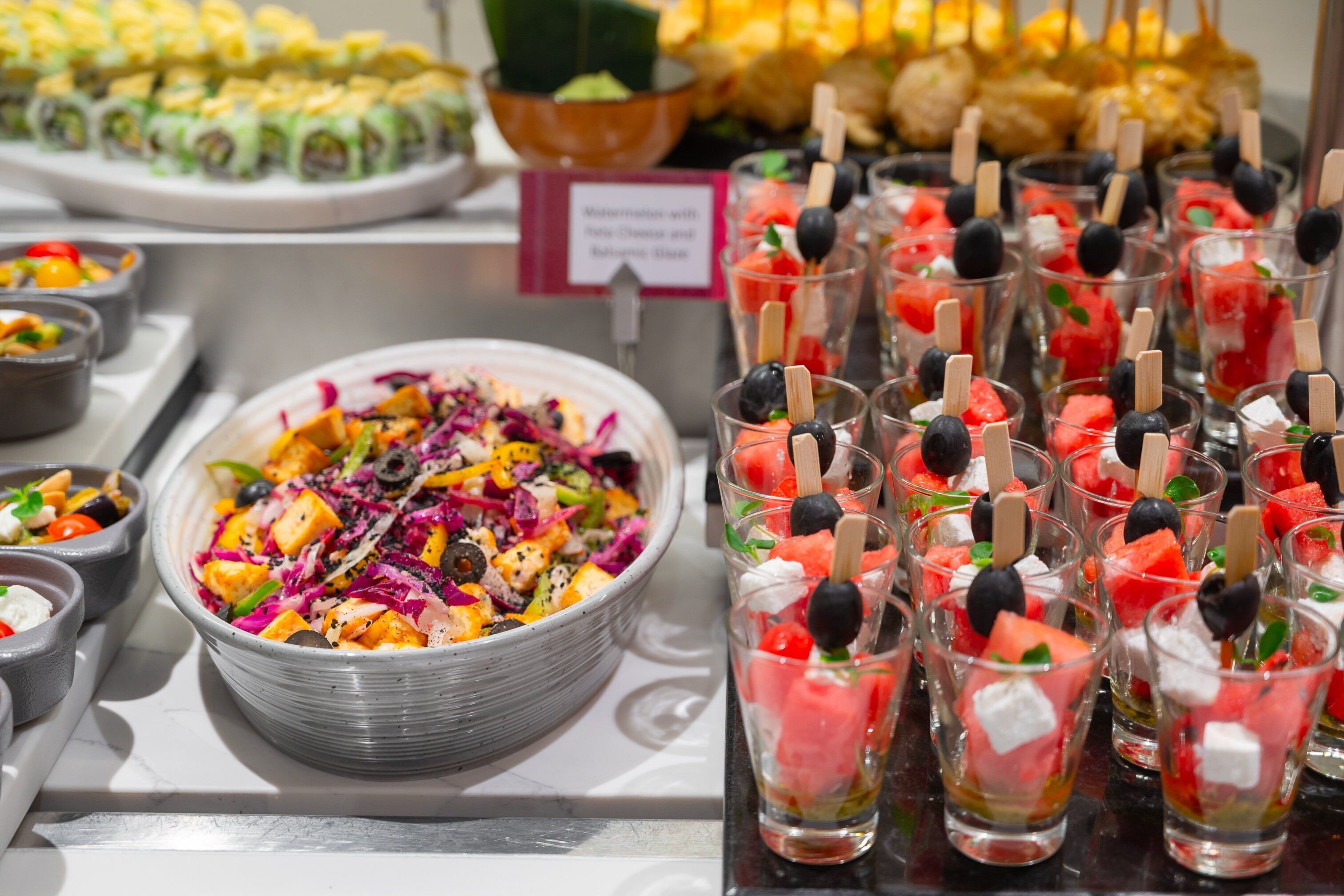 Un bufé con diversos alimentos, incluyendo una ensalada colorida, brochetas de sandía y otros platos.