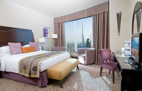 A hotel room with a large bed, two chairs, a desk with a TV, and a view of a city skyline.