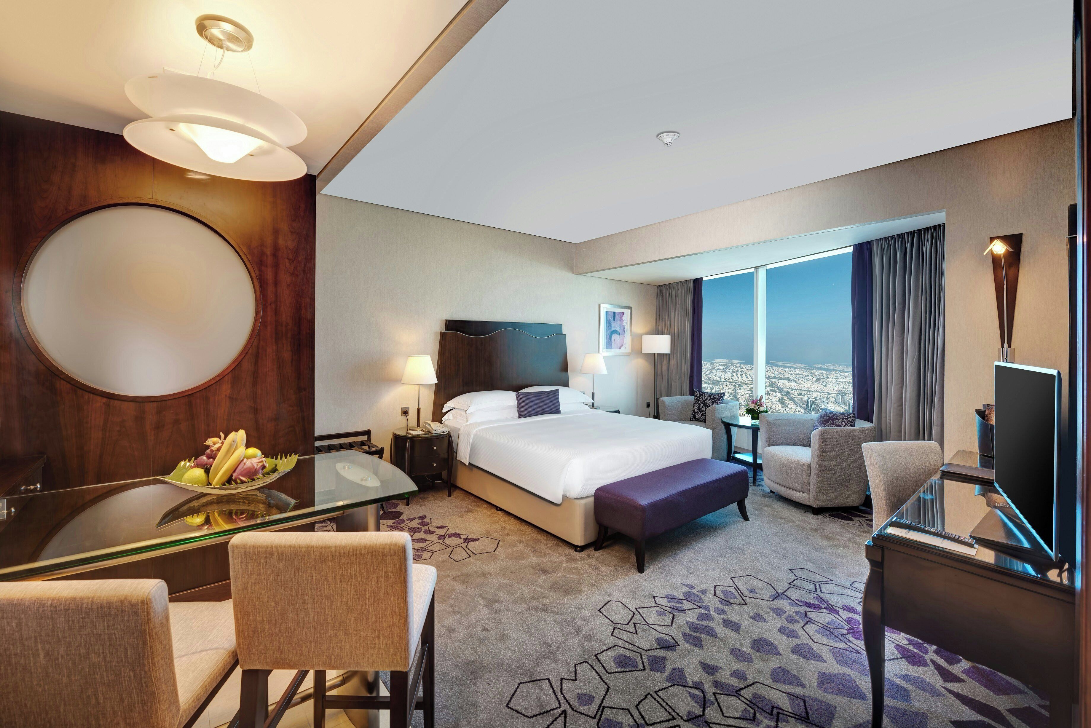 Habitación de hotel con cama, escritorio con silla, televisión y ventana grande con vista a la ciudad.