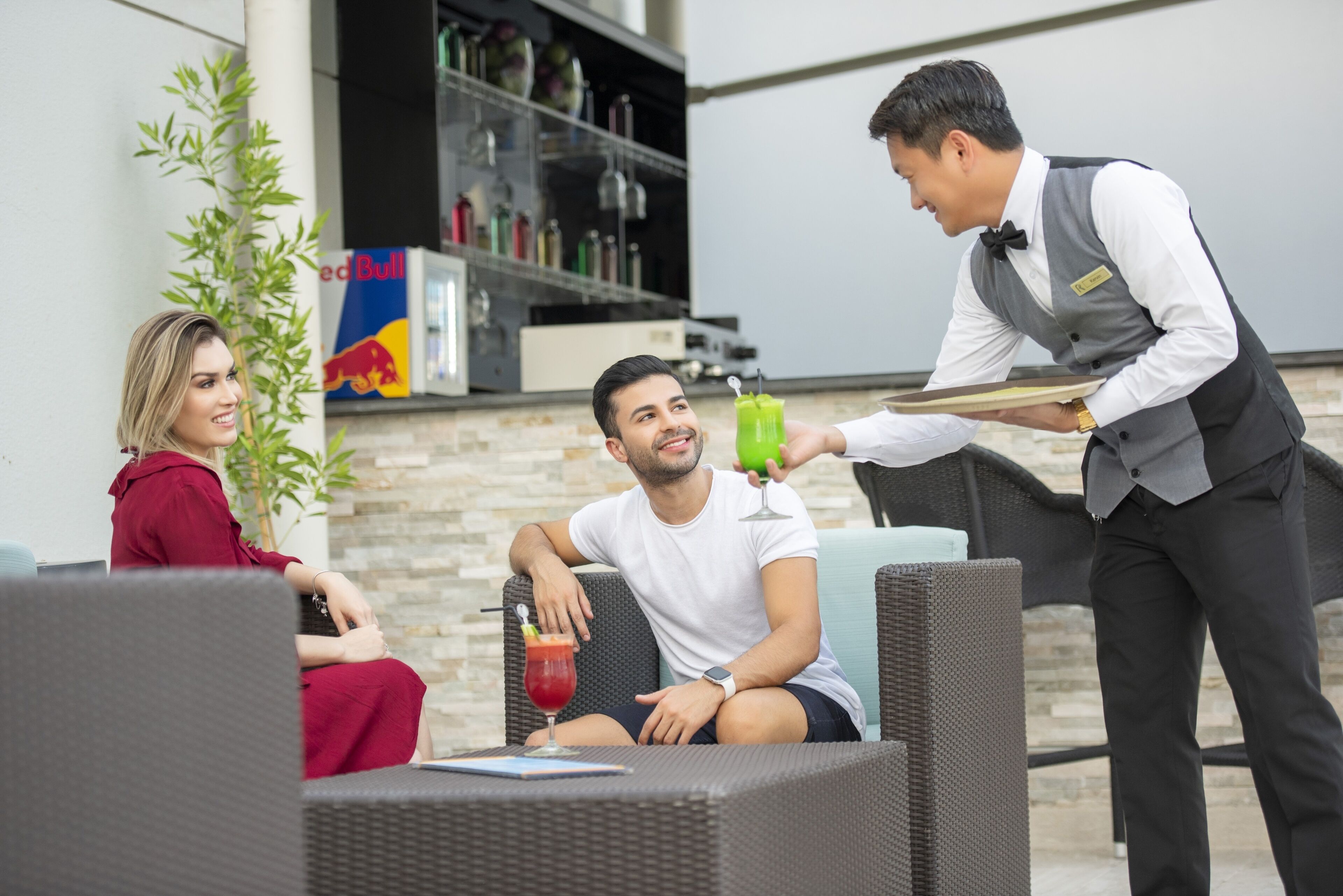 Une personne assise sur une chaise en osier, avec une boisson devant elle, une autre personne debout tenant un plateau, et un réfrigérateur Red Bull en arrière-plan.