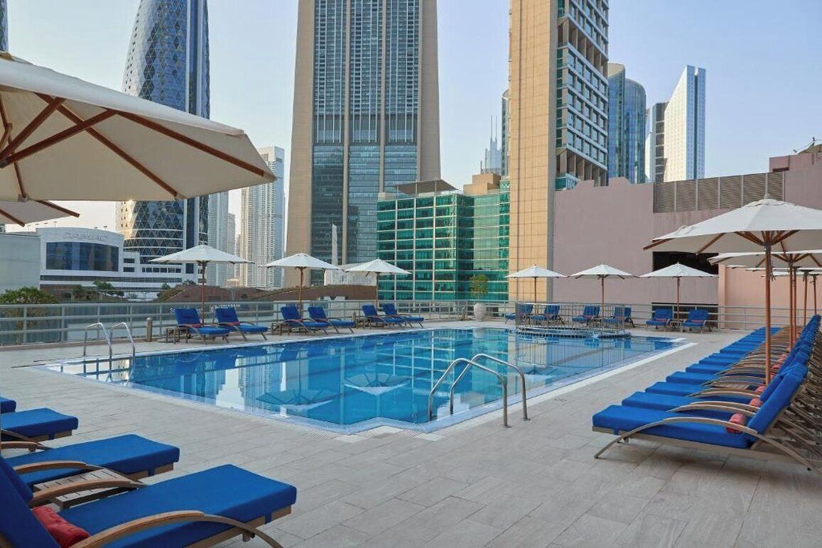 Una piscina en la azotea con sillones de descanso azules y sombrillas, rodeada de modernos rascacielos.