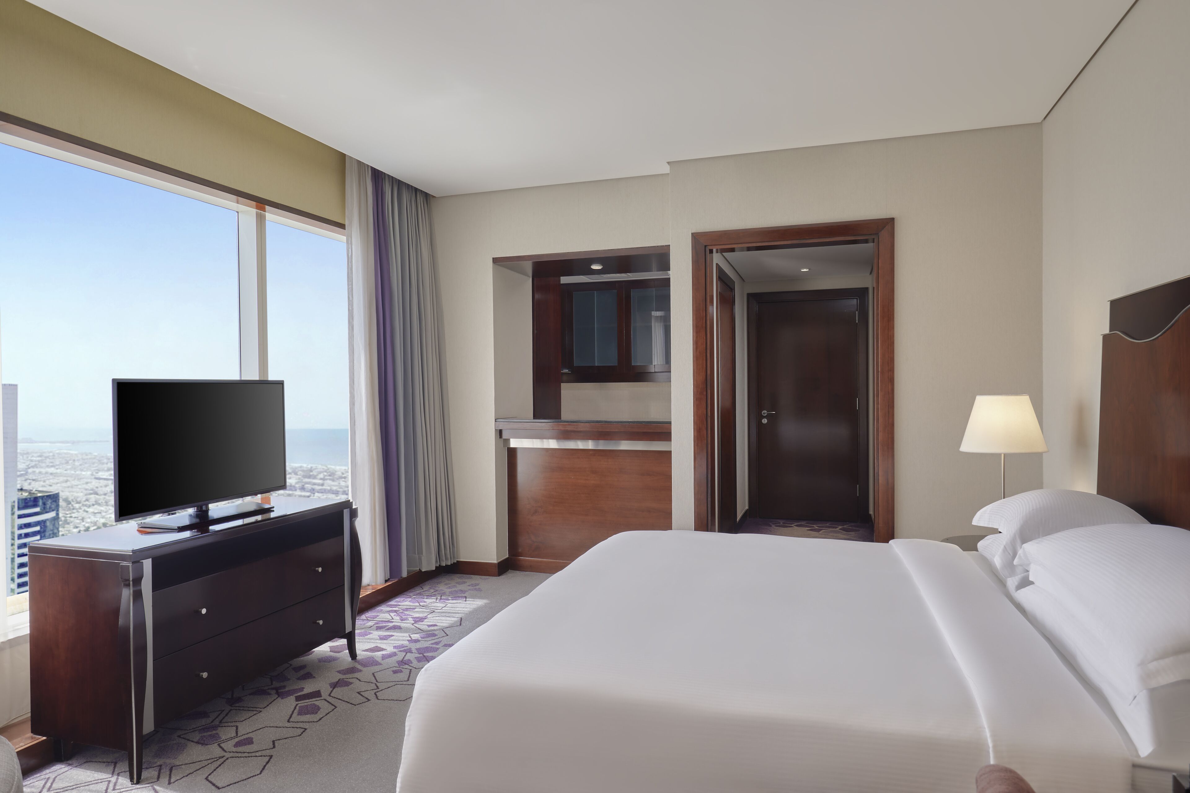 A hotel room with a large bed, a TV on a dresser, a desk, and a view of the city.