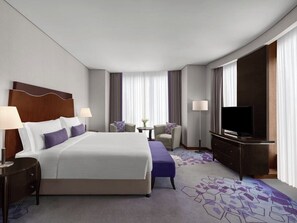 Habitación de hotel con una cama grande, dos mesitas de noche con lámparas, un televisor, una pequeña mesa redonda con una silla y un ventanal con cortinas.