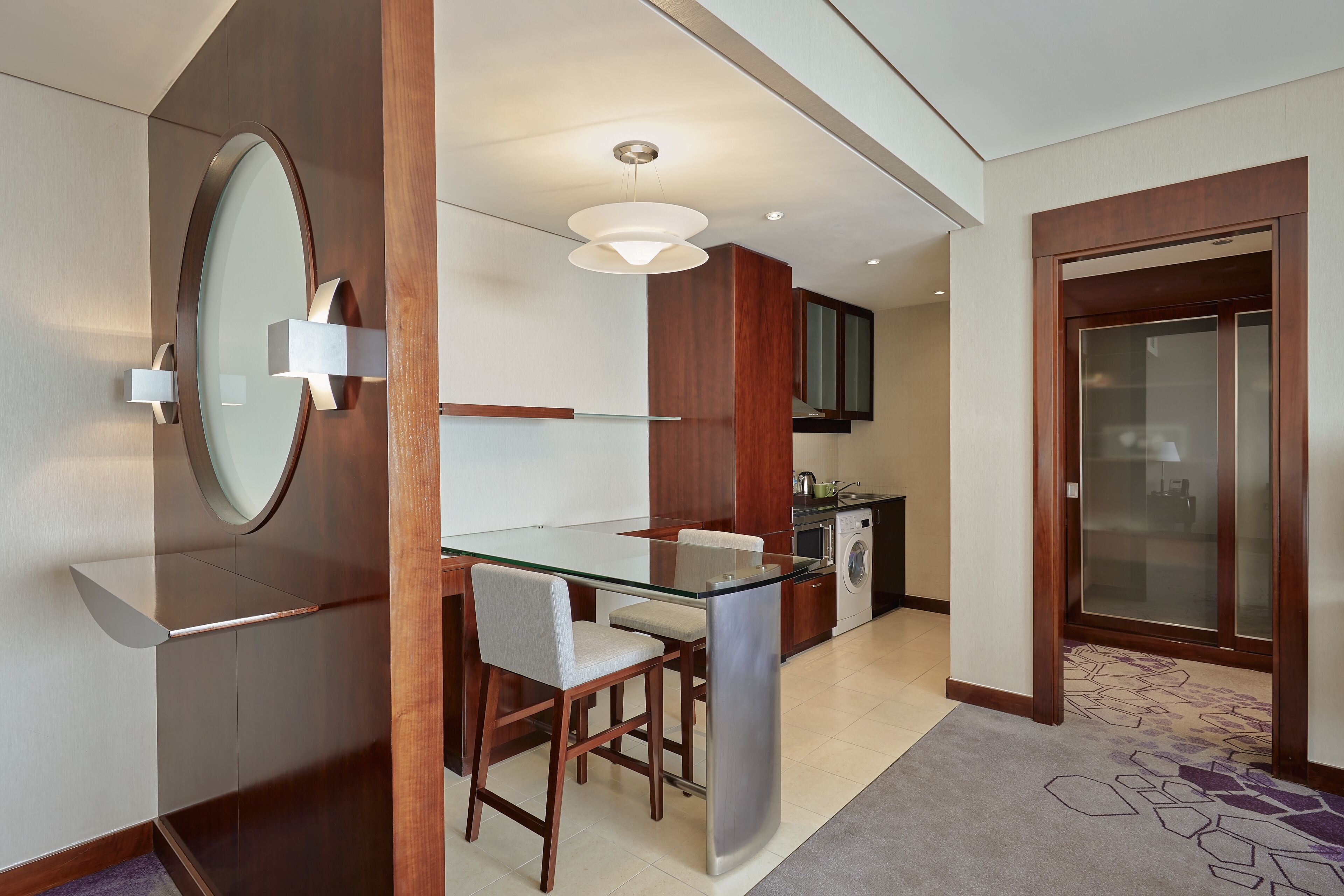 A modern hotel room with a glass dining table, a built-in kitchen, and a separate bathroom area.