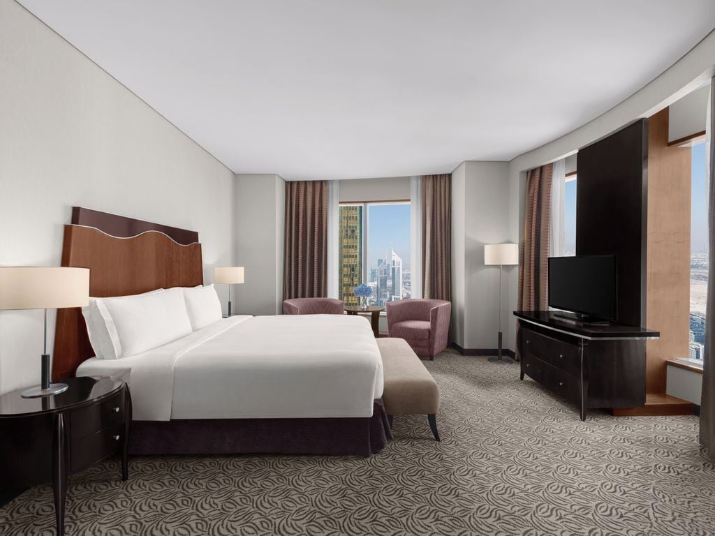 A hotel room with a bed, two chairs, a TV, and a view of a cityscape.