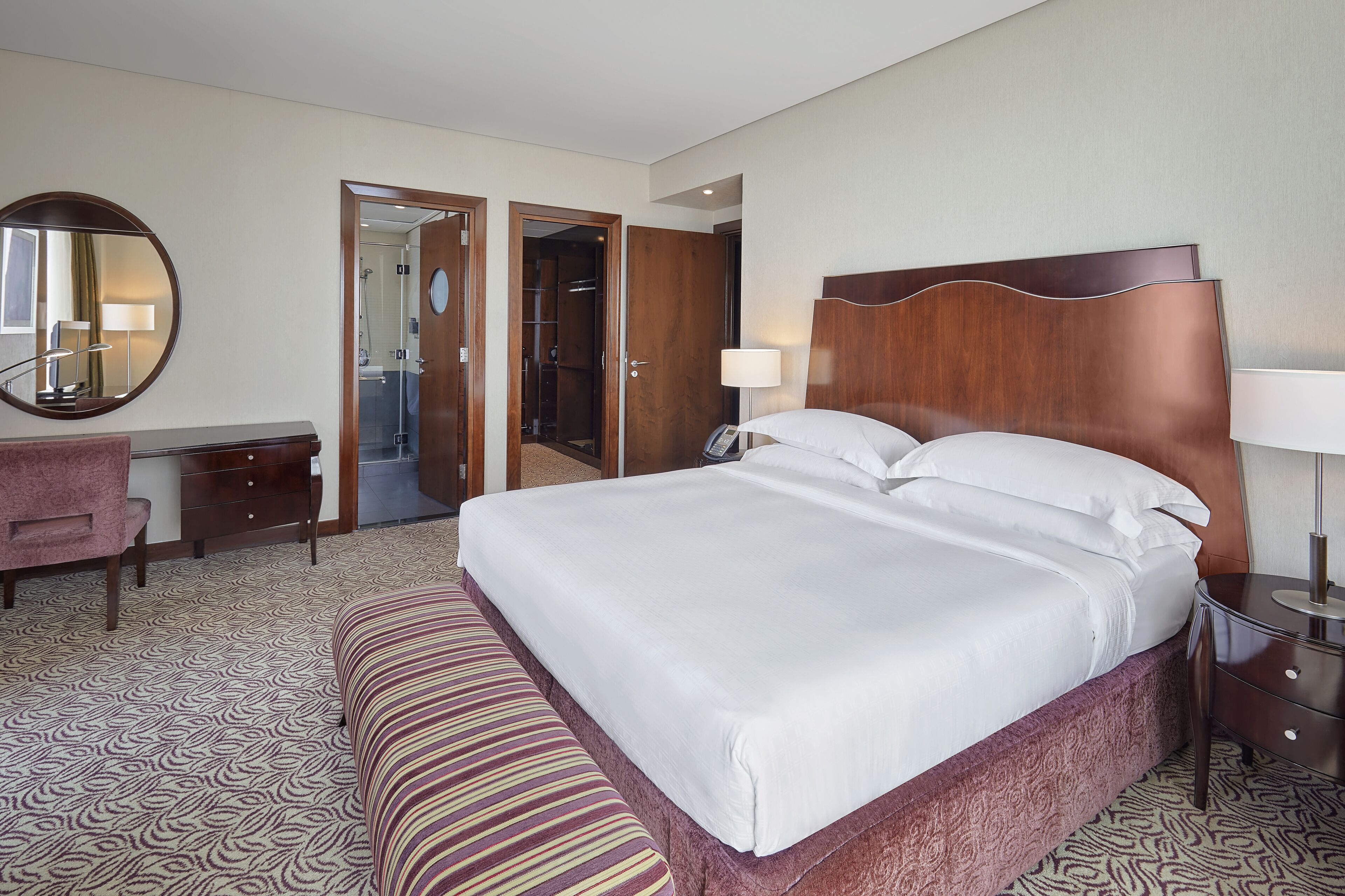 Ein Hotelzimmer mit einem großen Bett, einem Nachttisch, einem Schreibtisch, einem Sessel, einem Spiegel und einer offenen Tür zu einem Badezimmer.