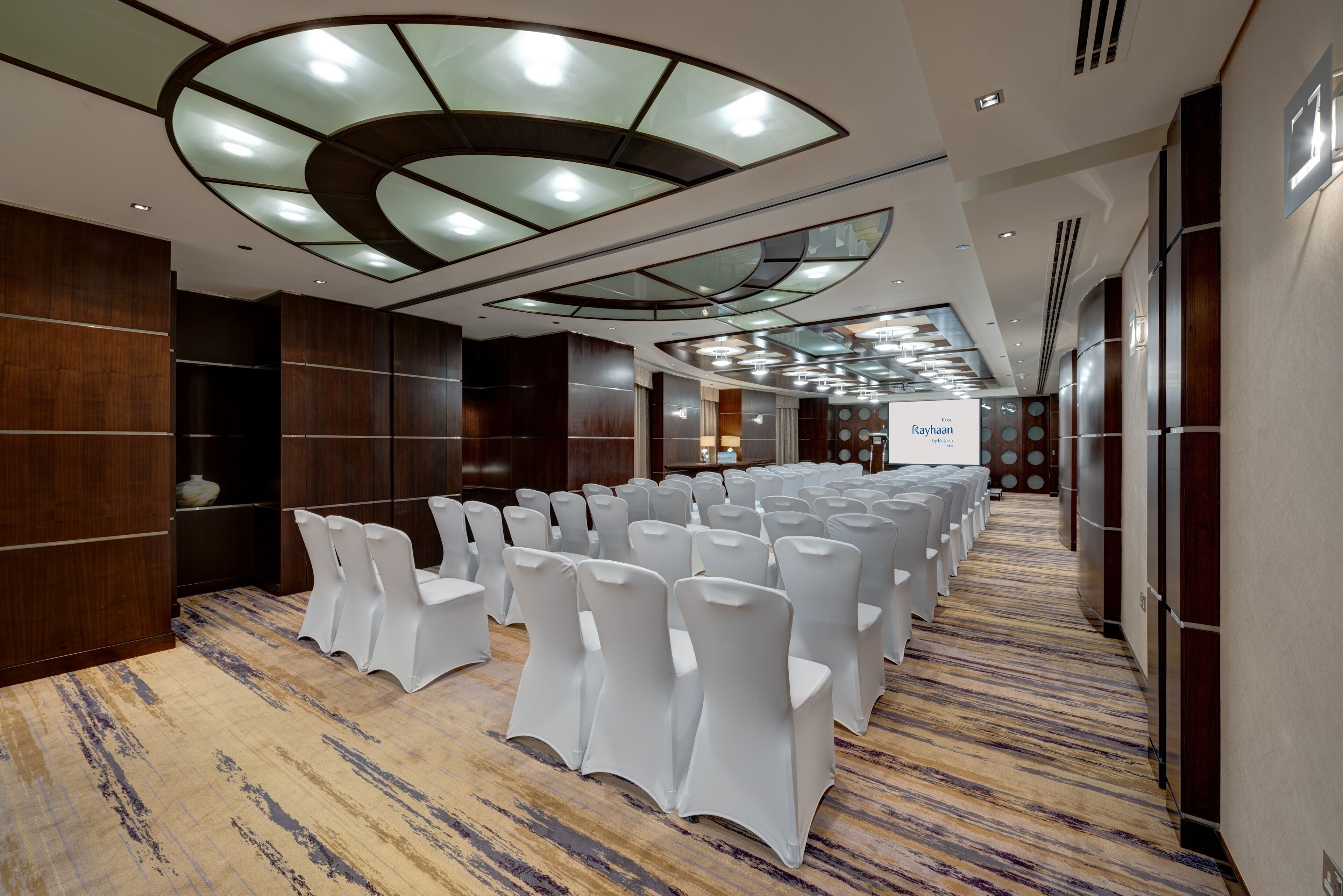 Une salle de conférence avec des rangées de chaises blanches, une moquette à motifs et un plafond au design moderne.