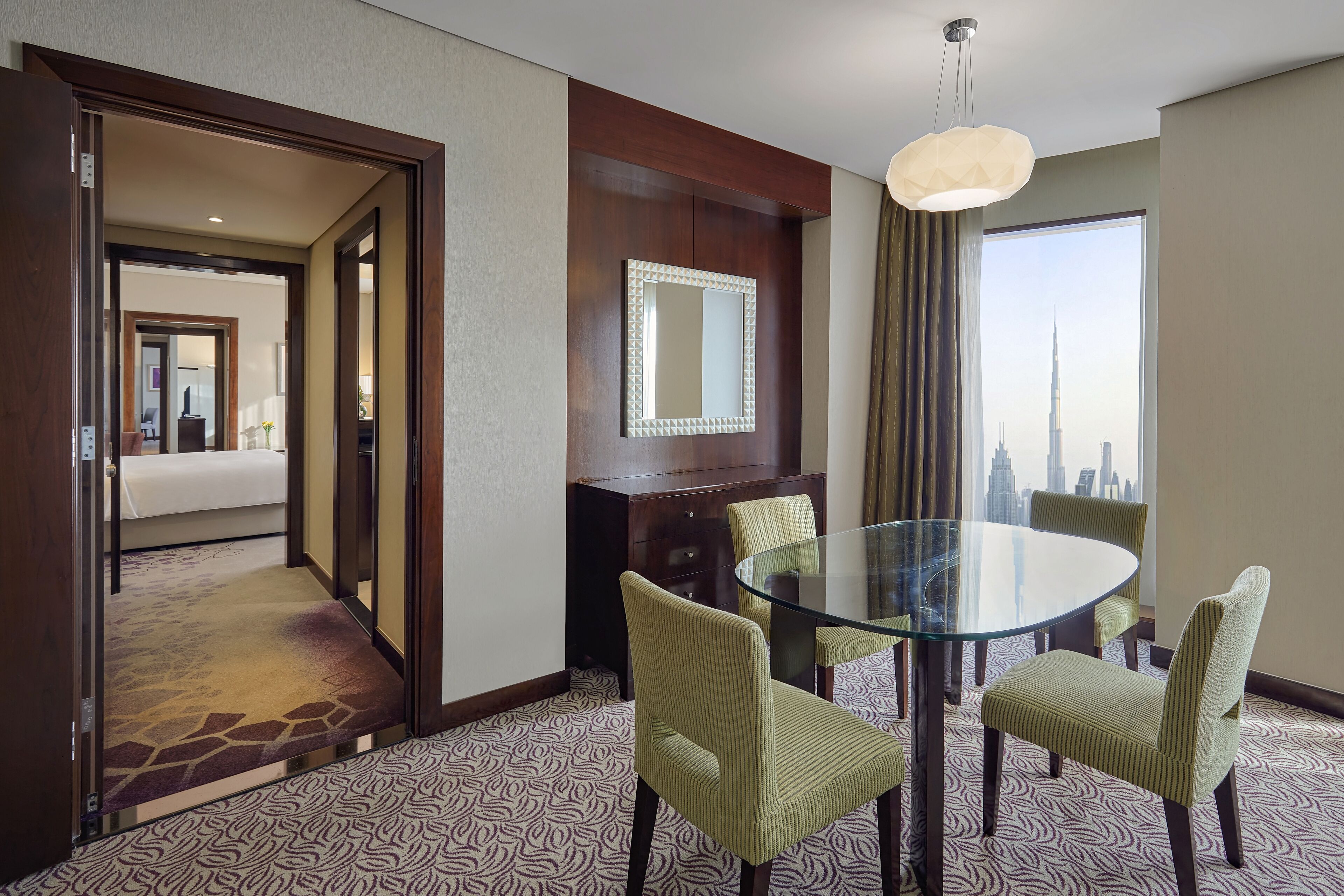 Ein Hotelzimmer mit Glastisch, vier Stühlen, einem Bett, einem Spiegel und Blick auf eine Stadtlandschaft.
