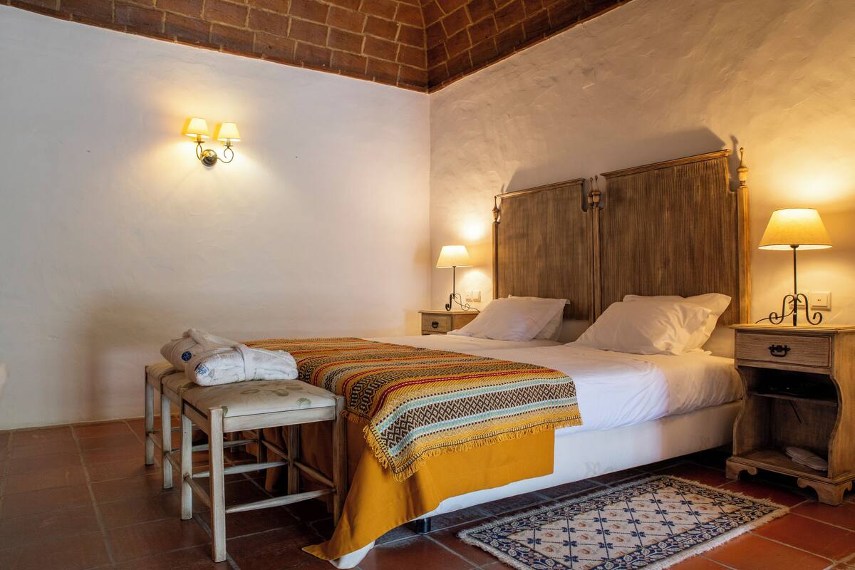 Un dormitorio con una cama, dos mesitas de noche, un banco y una alfombra estampada.