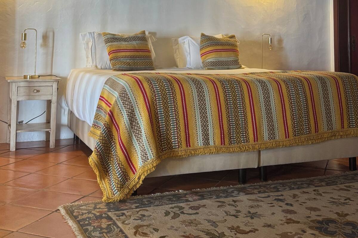 Una cama bien tendida con ropa de cama estampada, dos lámparas fijadas a la pared, una mesita de noche con una lámpara, y una alfombra estampada sobre un suelo de baldosas.