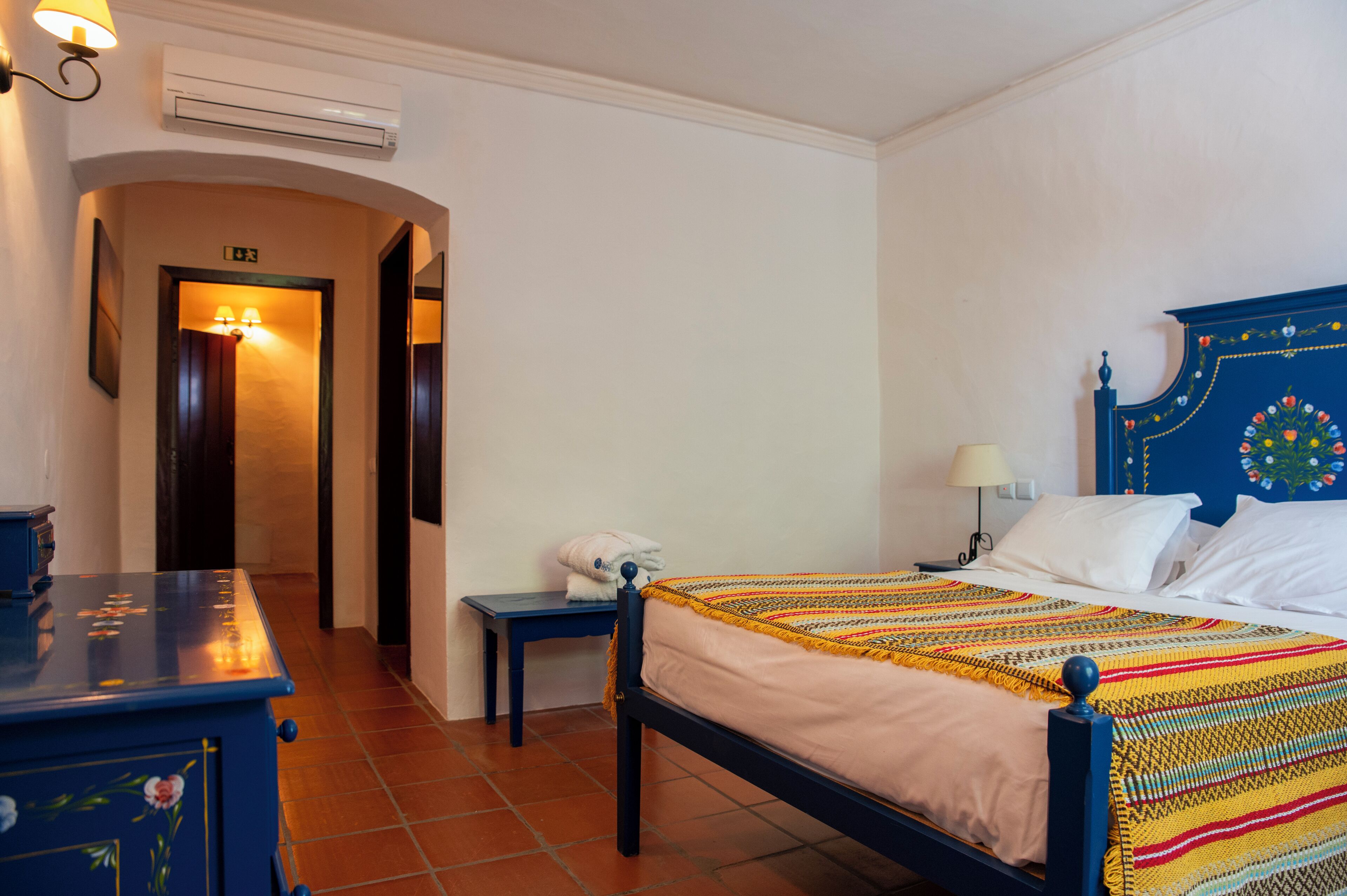 A hotel room with a bed, a nightstand, a dresser, and a hallway leading to an exit.