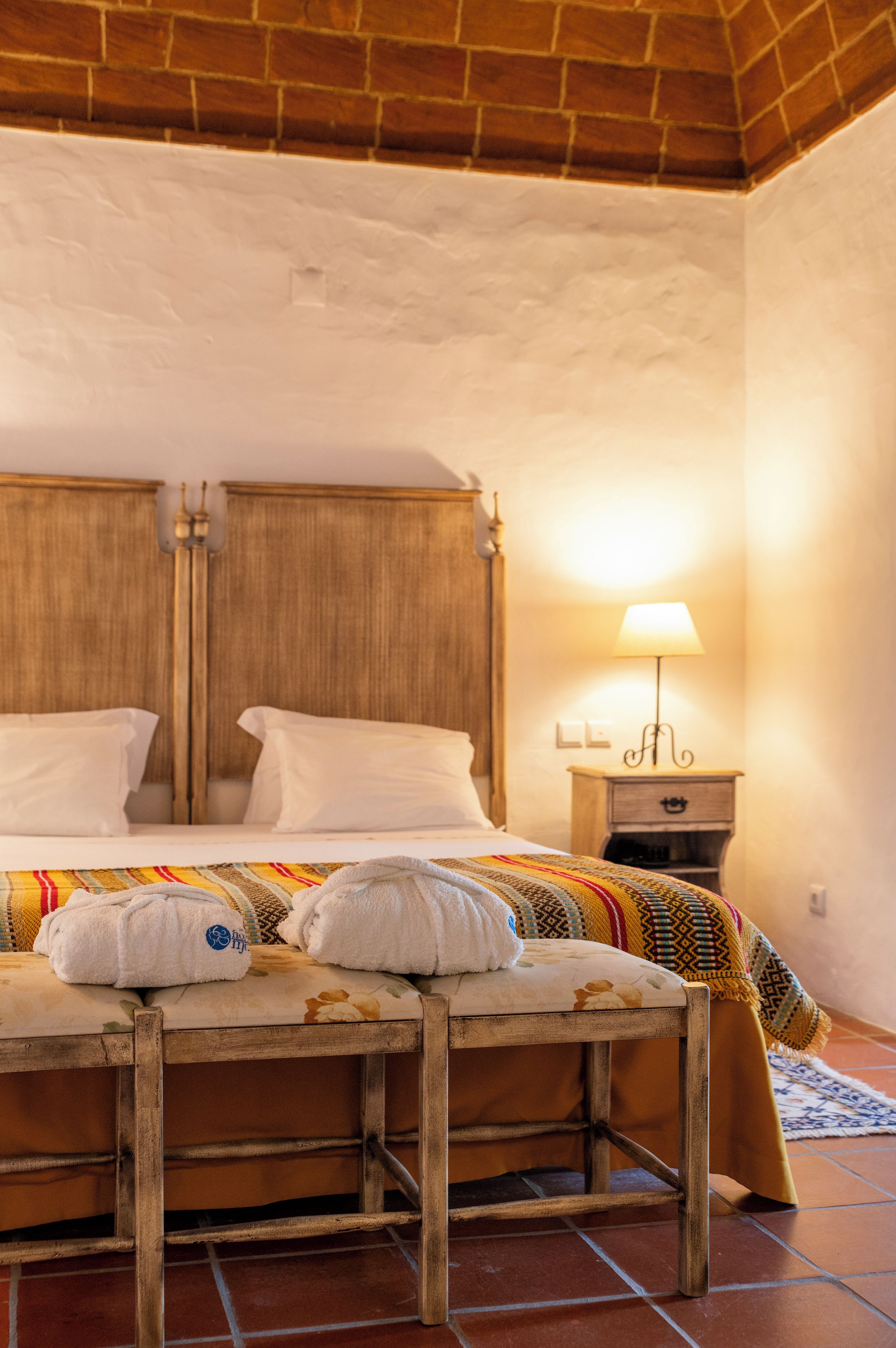 A hotel room with a wooden bed, white linens, a bedside table with a lamp, and a patterned rug.