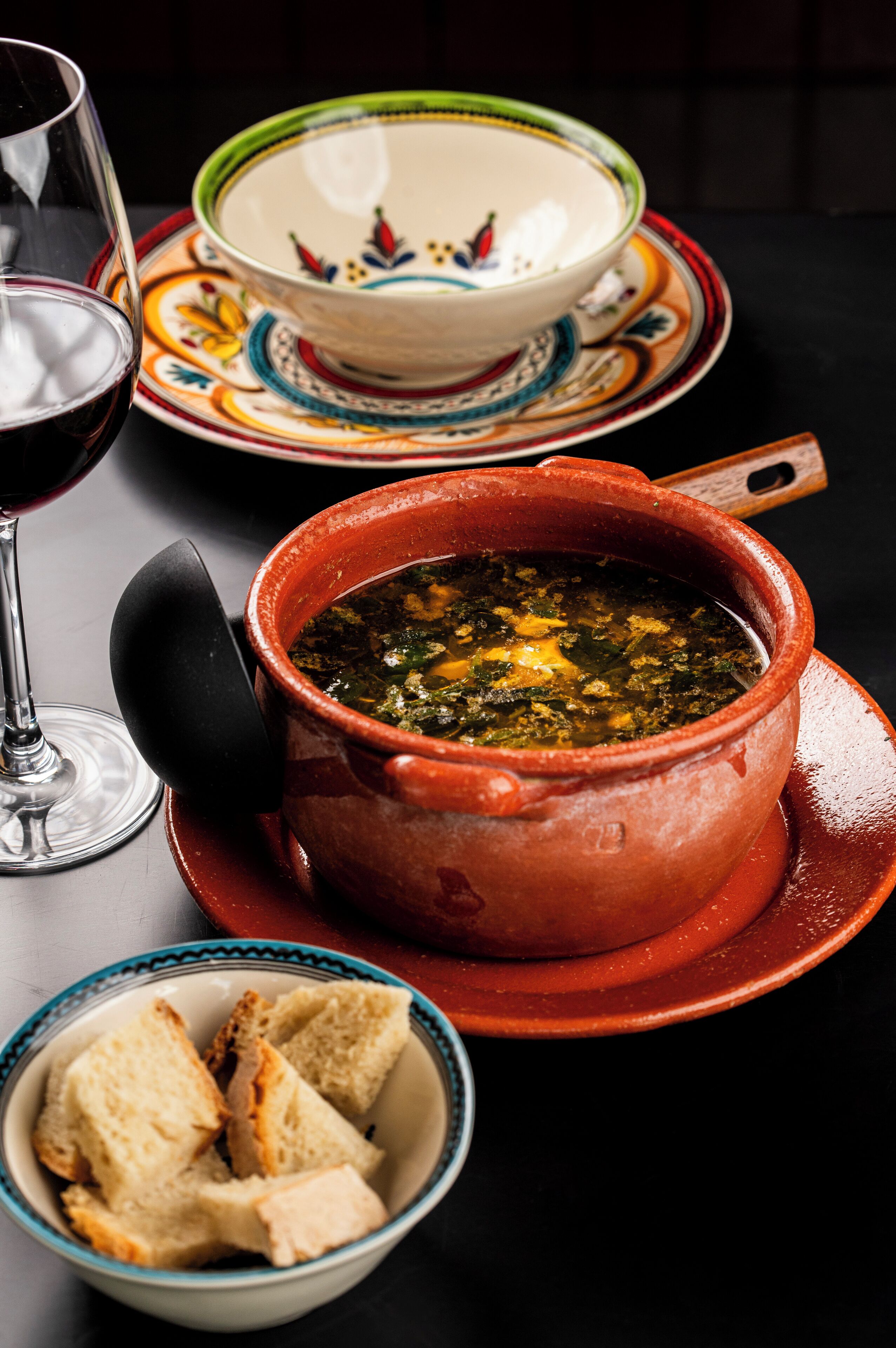 A bowl of soup with a lid, a glass of red wine, and a plate with bread slices.