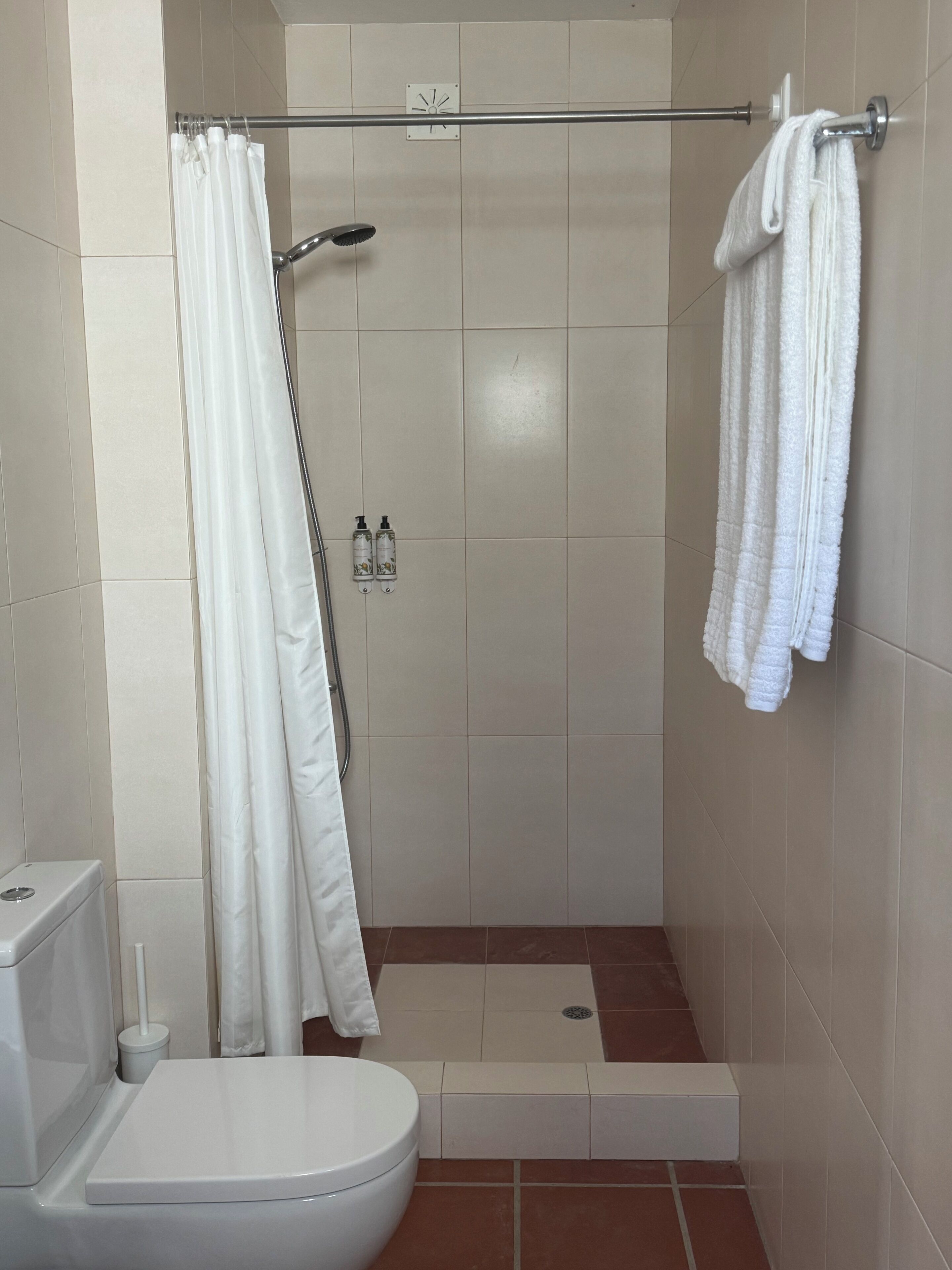 Een kleine, schone badkamer met toilet, douche en twee flessen shampoo.