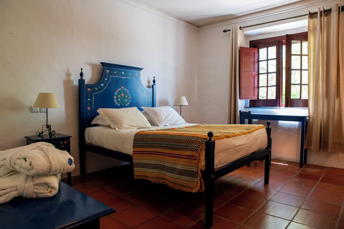 Una habitación con cama, mesita de noche, mesa y ventana con cortinas.