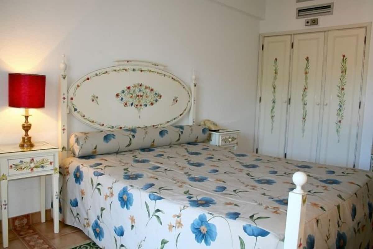 Un dormitorio con cubrecama estampado con flores, una mesita de noche con lámpara y un armario con motivos florales.