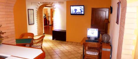 Un área de recepción con suelo de baldosas, mobiliario de mimbre, una computadora y un televisor.