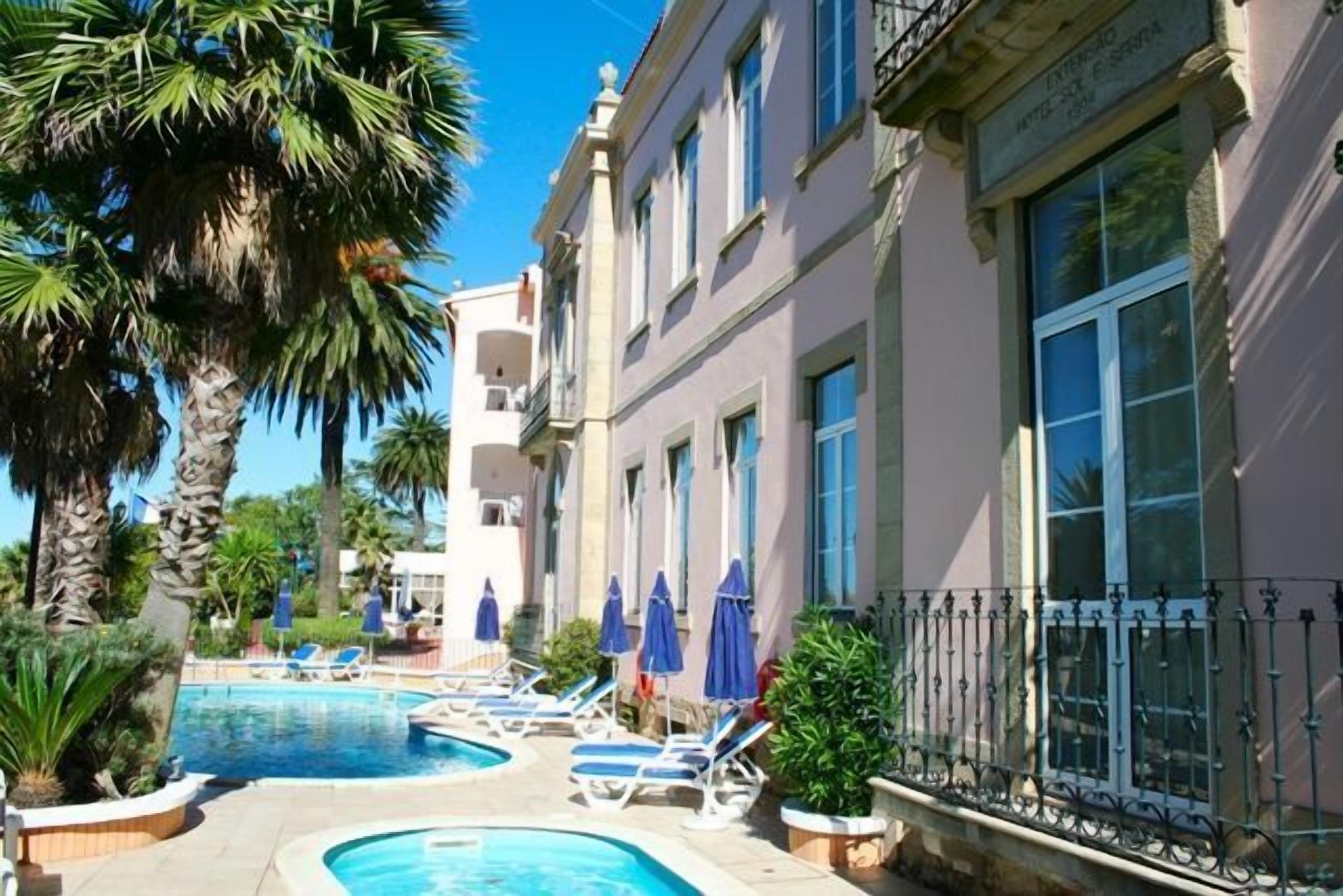 A pool area with lounge chairs and umbrellas, surrounded by palm trees and a building with balconies.