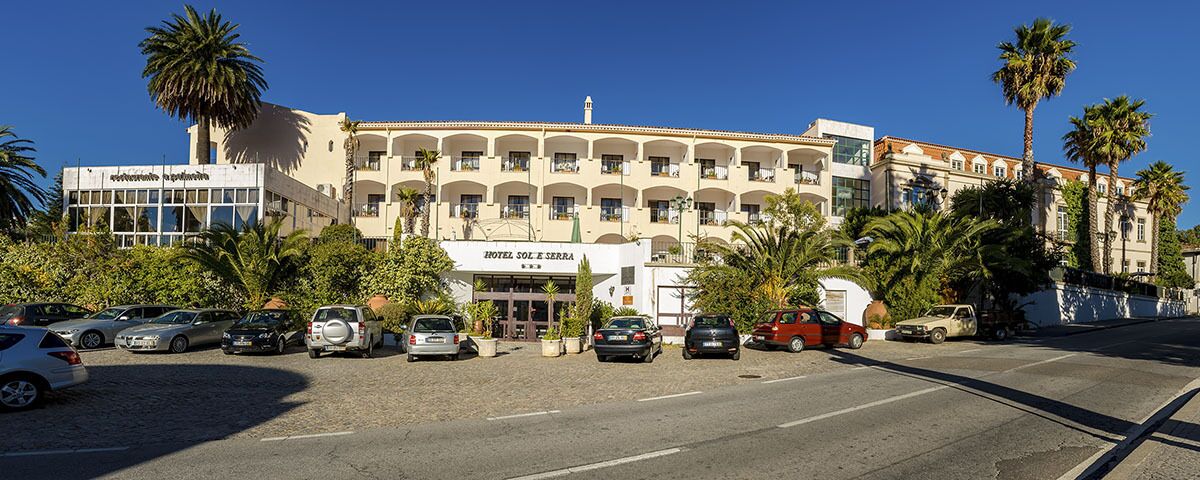 A hotel with a central entrance, multiple balconies, and a parking lot with several cars.