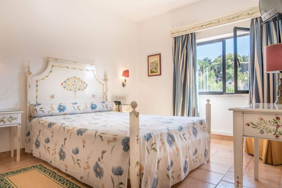 Un dormitorio con colcha estampada, una mesita de noche, una lámpara y una ventana con cortinas.