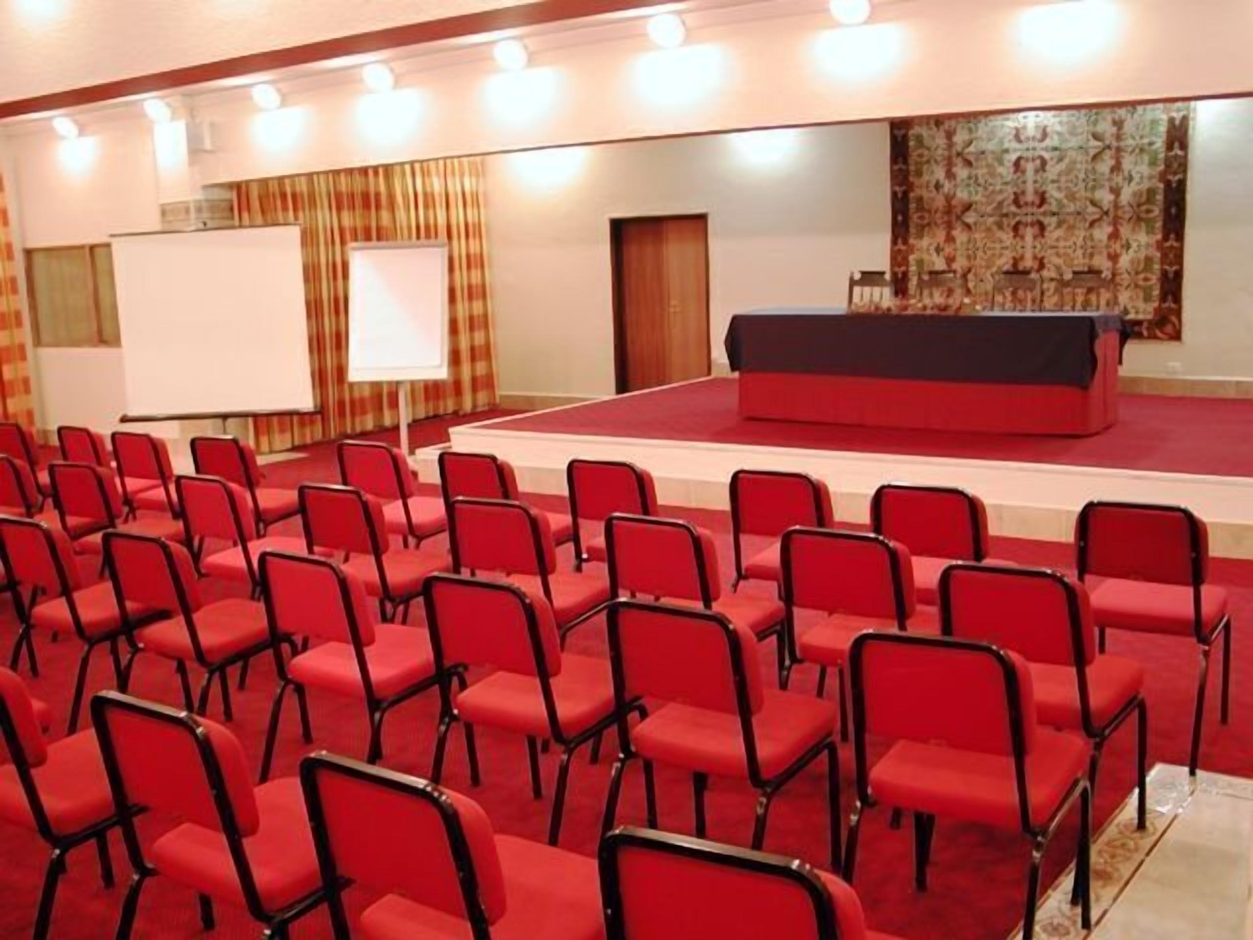 A conference room with rows of red chairs, a stage with a black table, and a whiteboard.