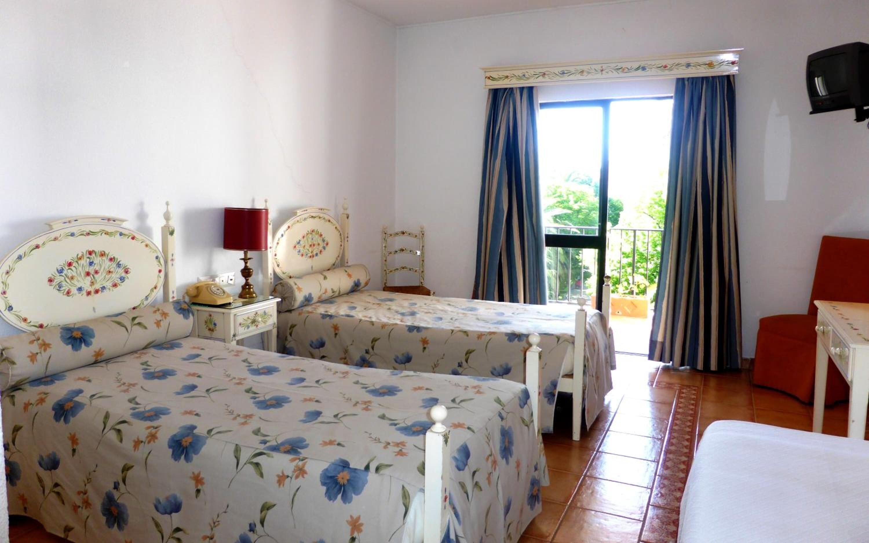 A room with two beds, a small table, a chair, a TV, and a balcony with curtains.