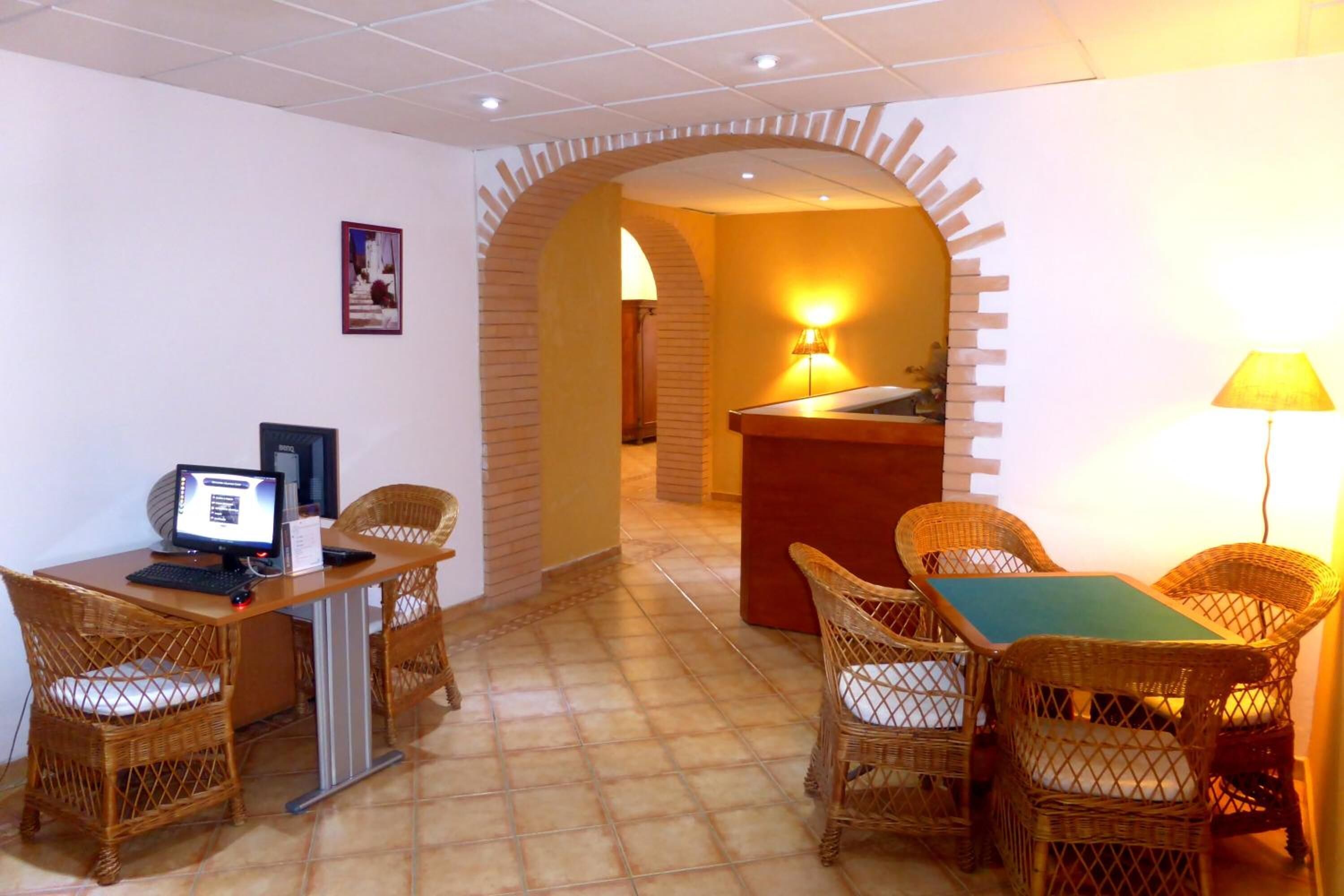 A hotel room with a computer desk, two wicker chairs, a small table, and a reception area with a lamp.