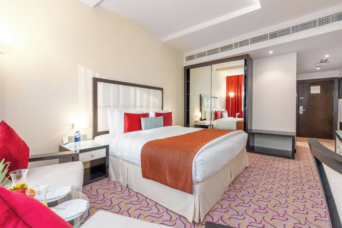 A hotel room with a large bed, a bedside table with a lamp, a chair, a small table with a glass, and a mirror.