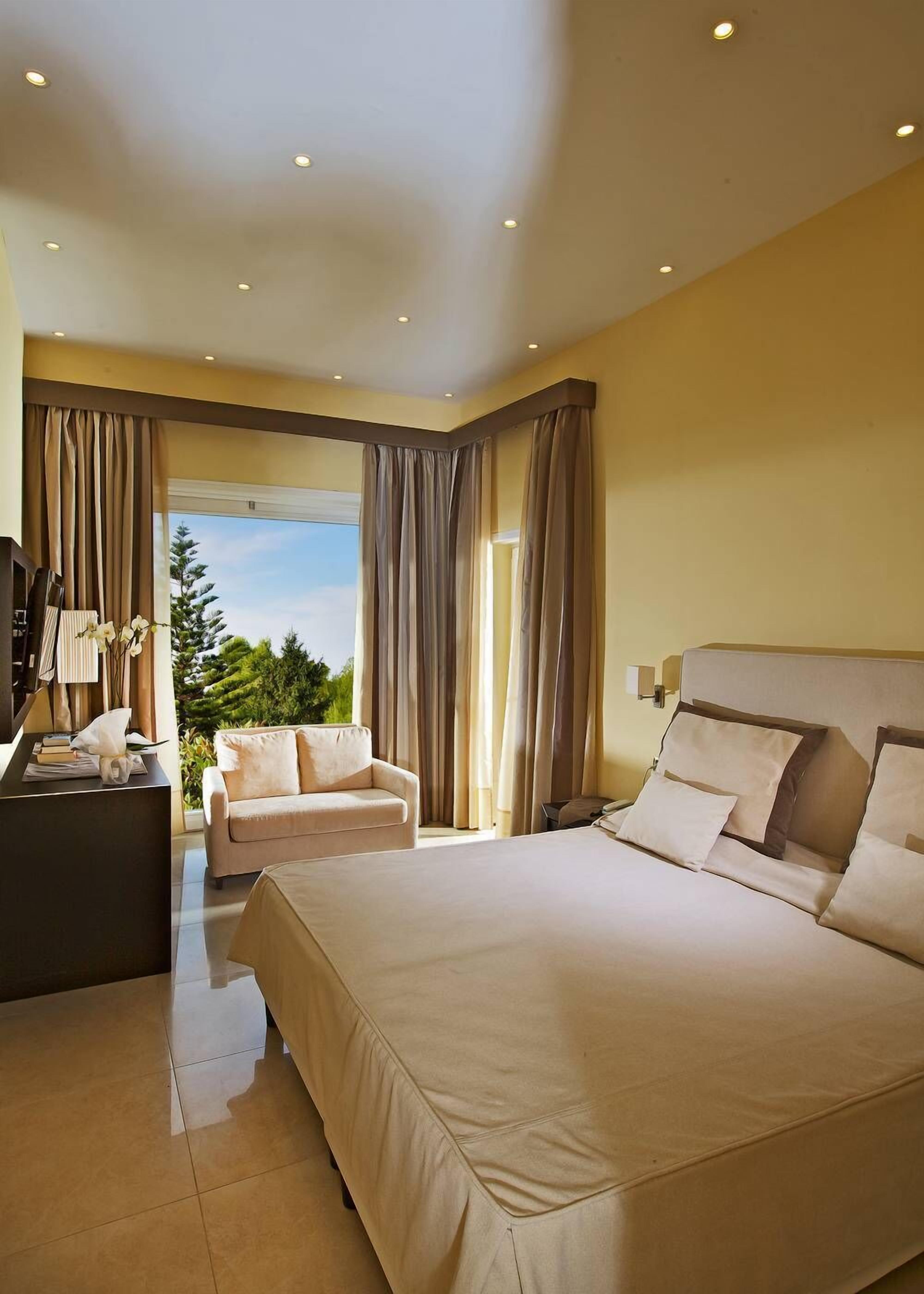 A modern hotel room with a large bed, a desk with a computer, a sofa, and a view of greenery outside.