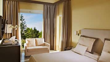 A modern hotel room with a large bed, a desk with a computer, a sofa, and a view of greenery outside.