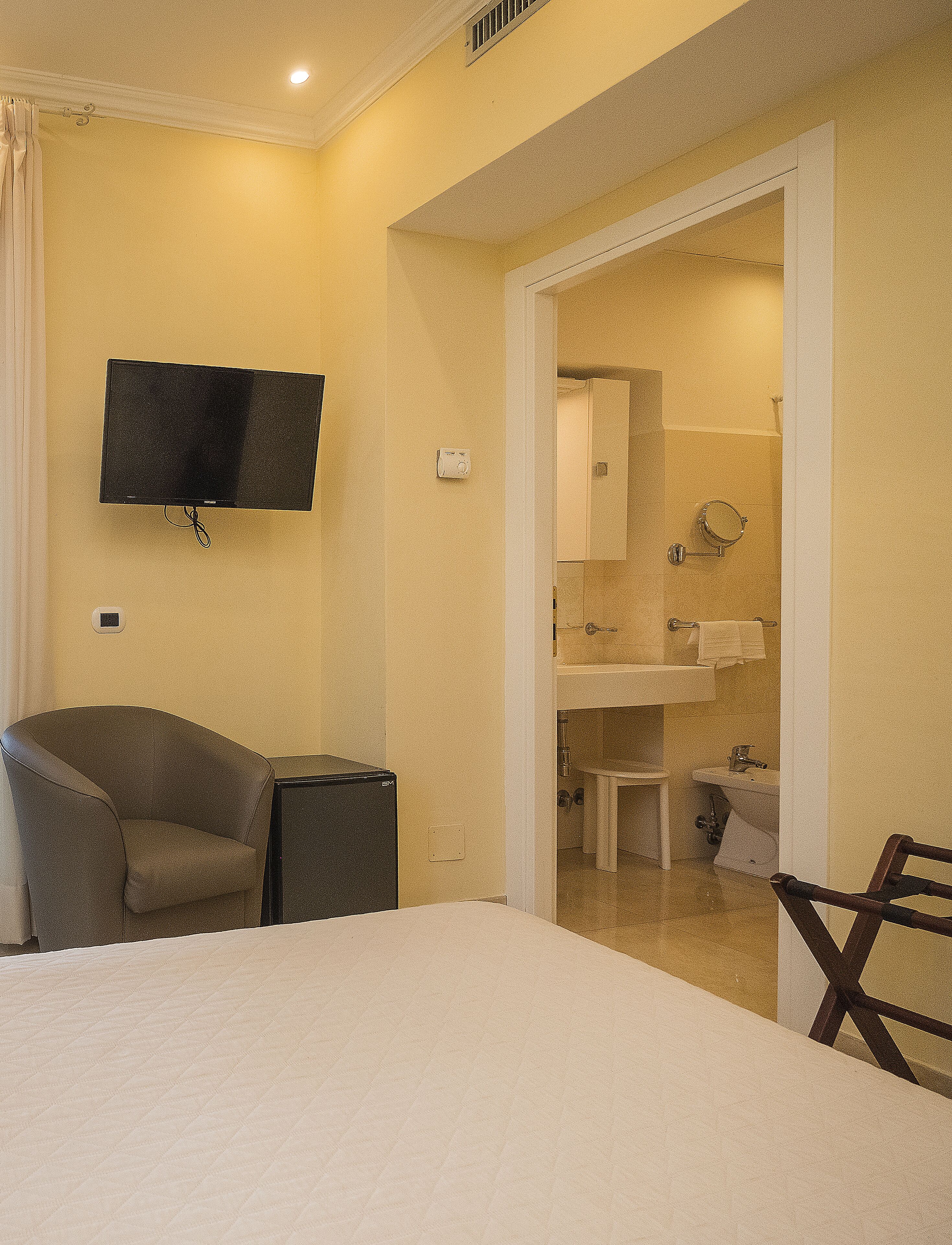 A hotel room with a bed, a TV mounted on the wall, a chair, a small table, and a bathroom visible through an open door.