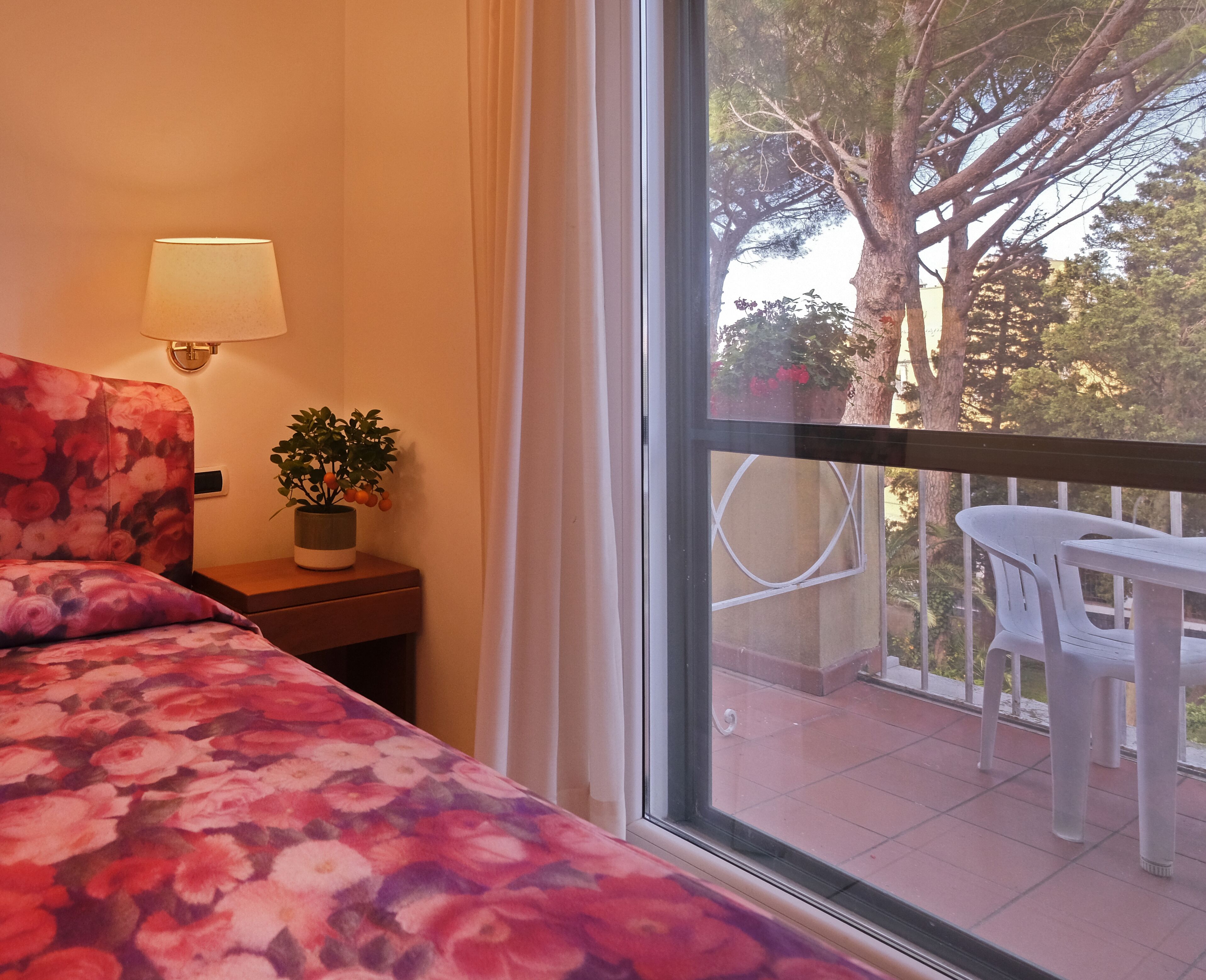 A hotel room with a bed, a nightstand with a lamp and a plant, and a balcony with a table and chairs.