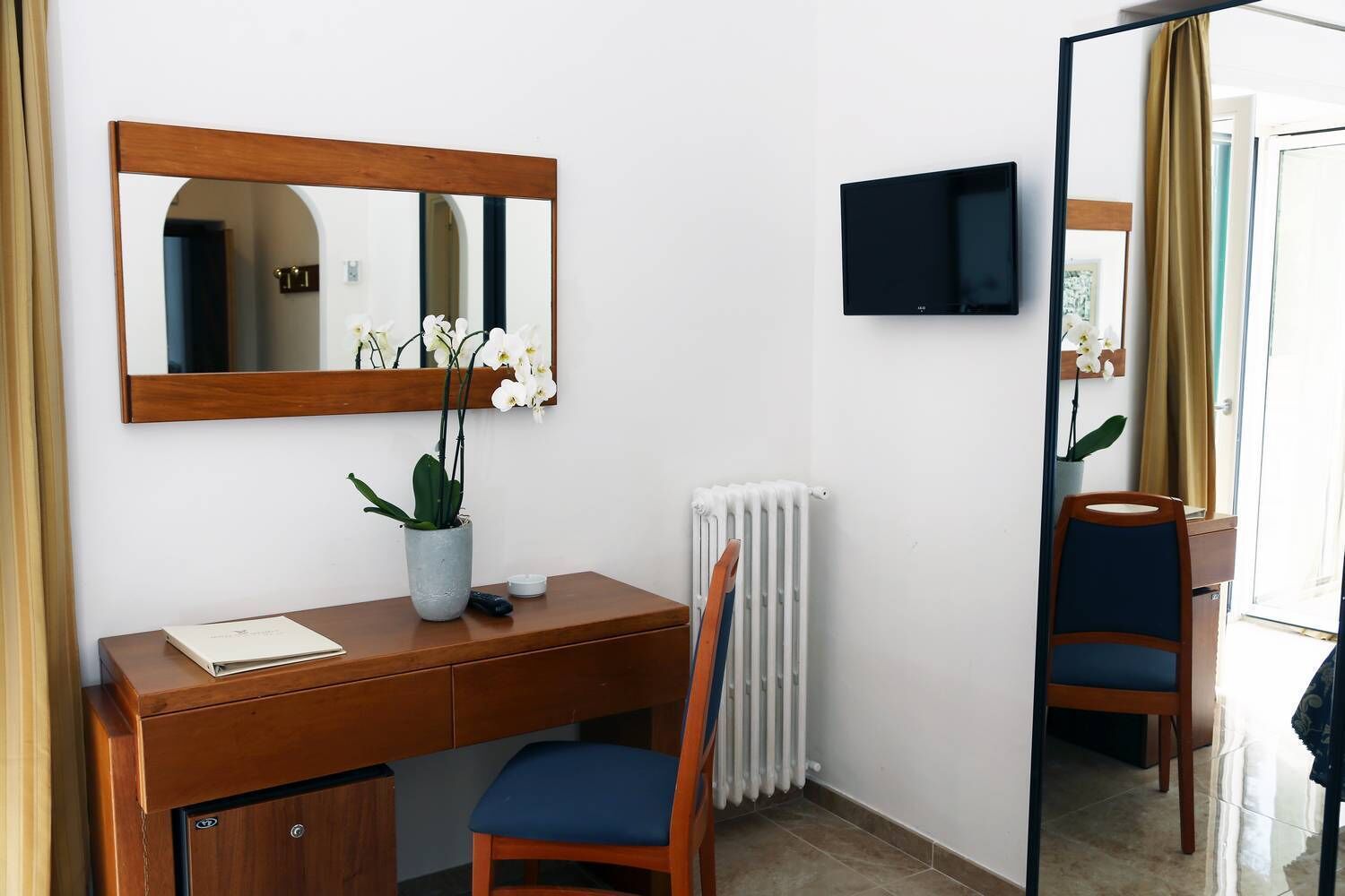 A hotel room with a wooden desk, a chair, a potted orchid, a radiator, and a television mounted on the wall.