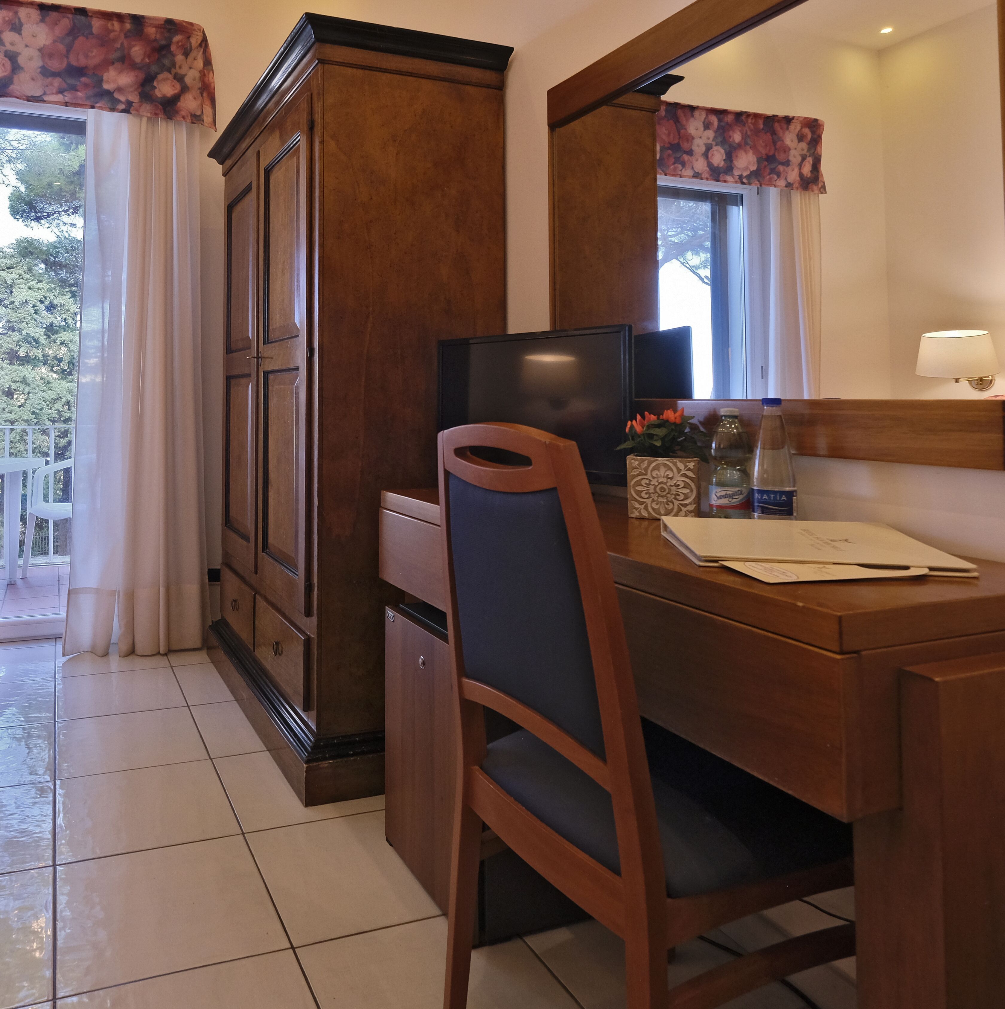 A wooden desk with a chair, a television, and a lamp. There is a cabinet with a floral curtain.